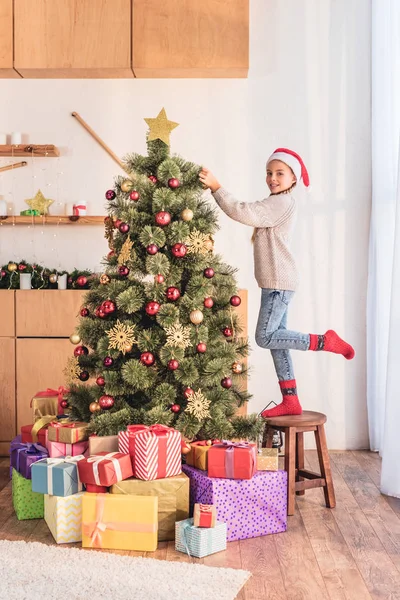 Smiling kid in santa hat standing on stool and decorating christmas tree at home with presents — Stock Photo