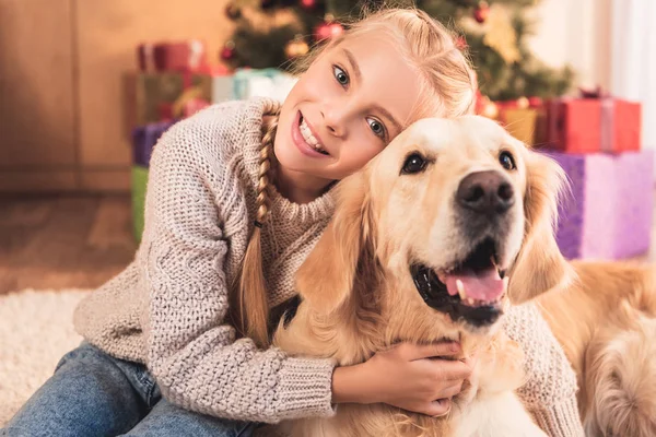 Lächelndes Kind umarmt Golden Retriever Hund und sitzt neben Weihnachtsbaum — Stockfoto
