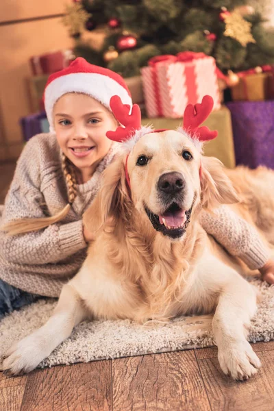 Улыбающийся ребенок в шляпе Санты и золотисто-ретриверской собаке с оленьими рогами, лежащими возле рождественских подарков — стоковое фото