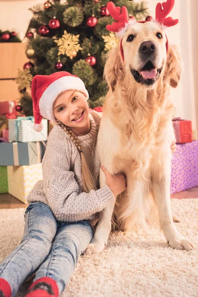 Garoto sorridente em chapéu de santa e cão golden retriever com chifres de veado sentado perto da árvore de natal com presentes — Fotografia de Stock