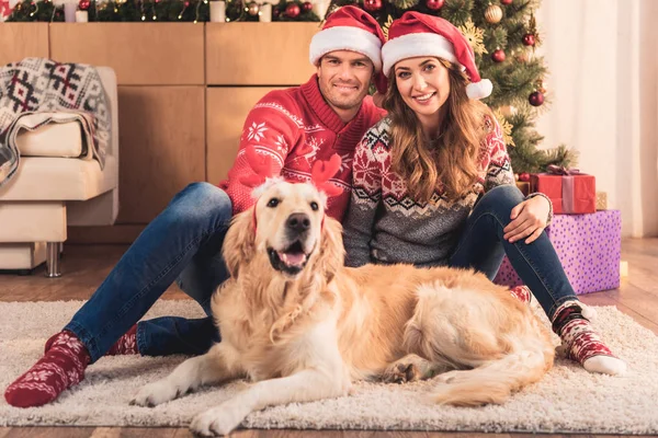 Schönes Paar in Weihnachtsmützen am Weihnachtsbaum sitzend mit Retriever-Hund in Hirschhörnern — Stockfoto