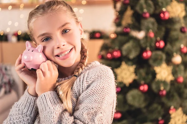 Glückliches Kind hält Sparschwein mit Weihnachtsbaum zu Hause — Stockfoto