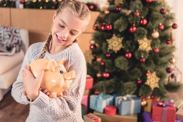 Adorable blonde child holding big piggy bank at home with christmas tree — Stock Photo