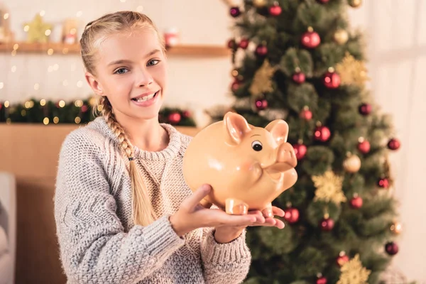 Smiling preteen kid holding big piggy bank at home with christmas tree — Stock Photo