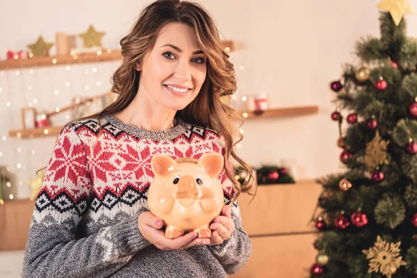 Bela mulher sorridente segurando porquinho banco com poupança em casa com árvore de natal — Fotografia de Stock