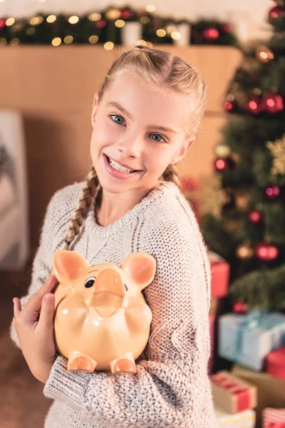 Adorable smiling kid holding piggy bank at home with christmas tree — Stock Photo