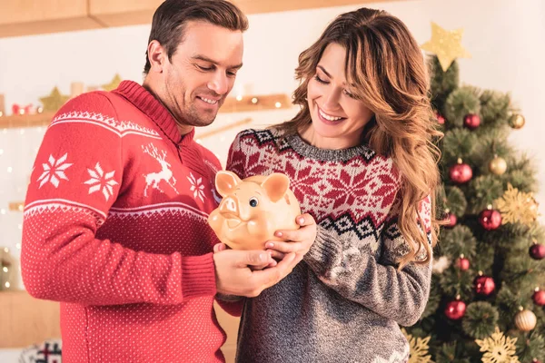 Улыбающаяся пара, держащая копилку со сбережениями дома с рождественской ёлкой — стоковое фото