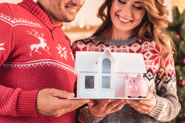 Couple in christmas sweaters holding house model and piggy bank, investment concept — Stock Photo