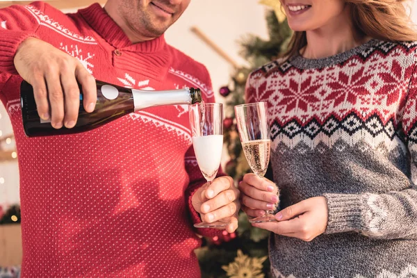 Cropped view of couple in christmas sweaters, husband pouring champagne for wife — Stock Photo