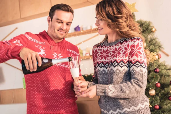 Casal feliz em camisolas de Natal, marido sorrindo derramando champanhe para a esposa — Fotografia de Stock