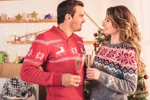 Smiling couple in christmas sweaters holding champagne glasses and looking at each other — Stock Photo