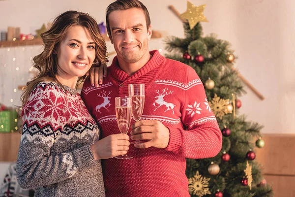 Sorrindo casal segurando copos de champanhe e olhando para a câmera em casa com árvore de natal — Fotografia de Stock