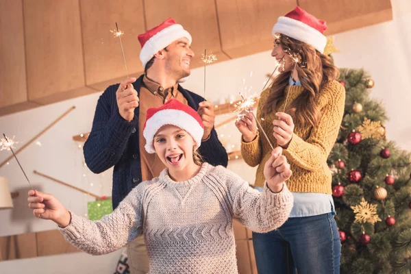 Família alegre em chapéus de santa comemorando com faíscas em casa com árvore de natal — Fotografia de Stock
