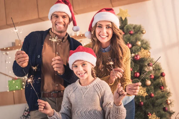 Sonrientes padres e hija en sombreros de santa celebración de bengalas en casa con árbol de Navidad - foto de stock