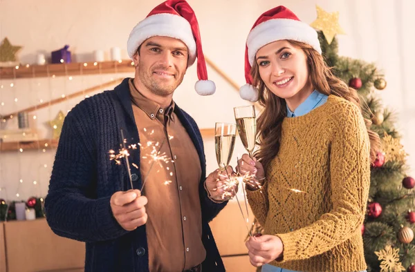 Casal feliz em chapéus de Papai Noel comemorando o Natal com copos de champanhe e sparklers — Fotografia de Stock