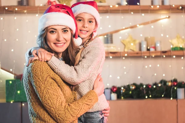 Mère et fille souriantes dans des chapeaux de Père Noël étreignant à la maison au moment de Noël — Photo de stock