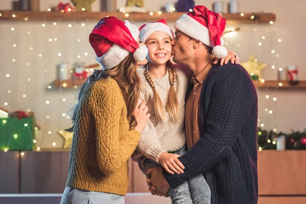 Счастливые родители в шляпах Санты целуют улыбающуюся дочь на Рождество — стоковое фото