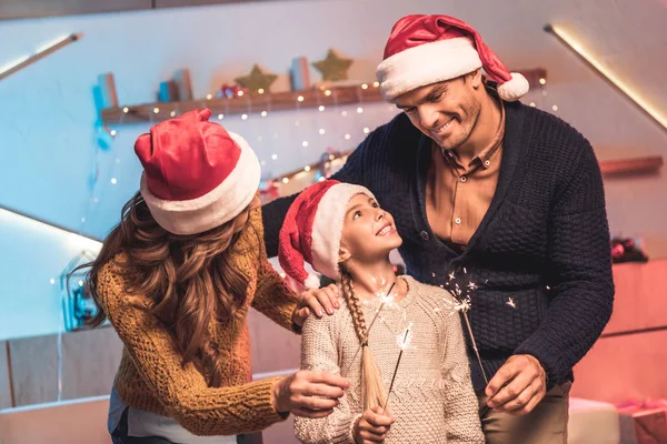 Padres felices e hija en sombreros de santa celebrando la Navidad con bengalas - foto de stock