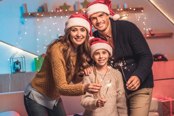Familia sonriente en sombreros de santa celebrando el año nuevo con bengalas - foto de stock