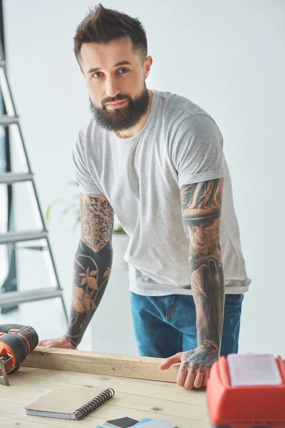 Красивий бородатий чоловік спирається на дерев'яний стіл і дивиться на камеру під час ремонту будинку — стокове фото