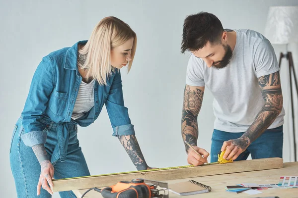 Jeune couple tatoué travaillant avec crayon, planche en bois et ruban à mesurer — Photo de stock
