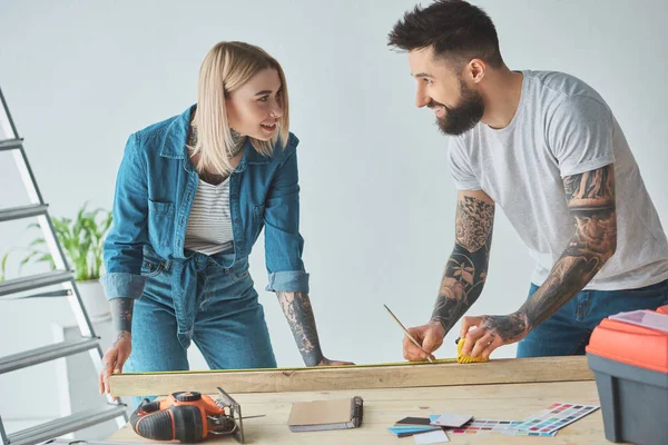 Joven pareja tatuada sonriendo entre sí mientras sostiene lápiz, tablón de madera y cinta métrica - foto de stock