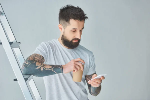 Bearded young man holding paper cup and using smartphone while standing near stepladder on grey — Stock Photo