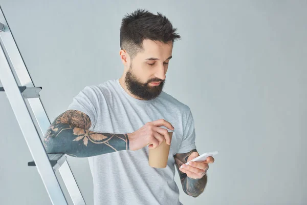 Joven barbudo sosteniendo una taza de papel y usando un teléfono inteligente mientras está parado cerca de la escalera en gris - foto de stock