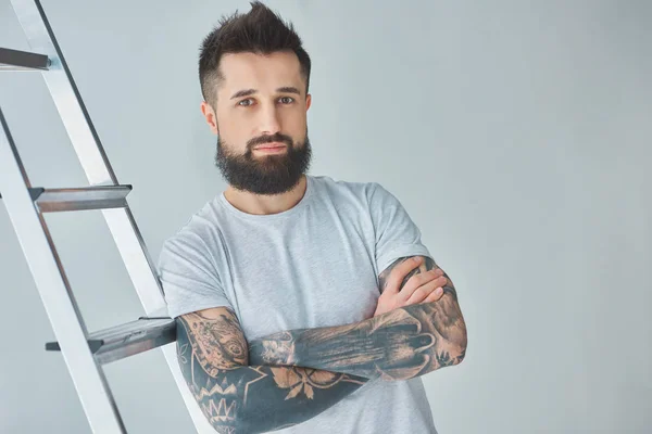 Handsome bearded tattooed young man standing with crossed arms near stepladder and looking at camera on grey — Stock Photo