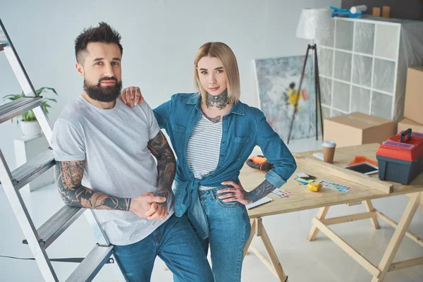 Schönes junges tätowiertes Paar blickt in die Kamera, während es im neuen Zuhause zusammen steht — Stockfoto