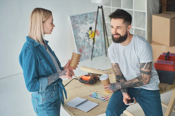 Happy young couple drinking coffee from paper cups and using smartphone during home improvement — Stock Photo