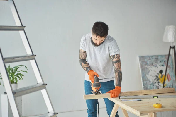 Handsome bearded tattooed man using electric jigsaw during house repair — Stock Photo
