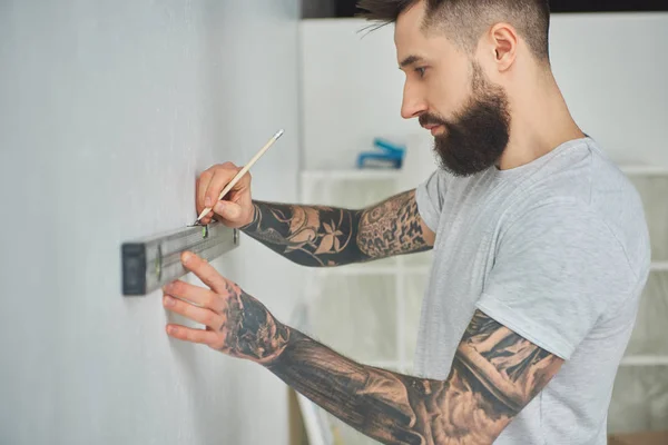 Visão lateral de belo jovem barbudo segurando ferramenta de nível e lápis durante a melhoria em casa — Fotografia de Stock