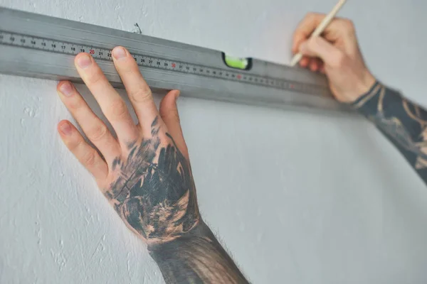 Cropped shot of tattooed man holding level tool and marking wall with pencil during repairs — Stock Photo