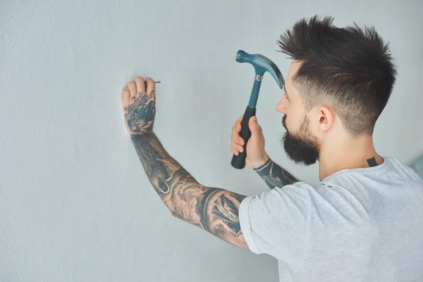 Bearded tattooed young man hammering nail at wall — Stock Photo