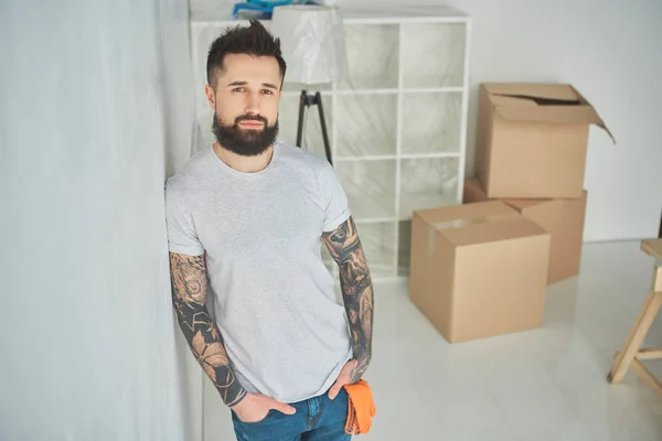 Красивый бородатый татуированный мужчина, склоняющийся к стене и смотрящий в камеру в новом доме — стоковое фото