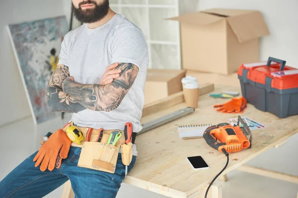 Schnappschuss eines bärtigen tätowierten Mannes mit Werkzeuggürtel, der mit verschränkten Armen auf einem Holztisch sitzt — Stockfoto