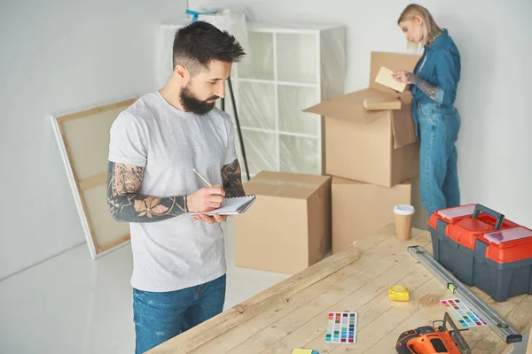 Bearded man taking notes and young woman unpacking cardboard boxes in new house — Stock Photo