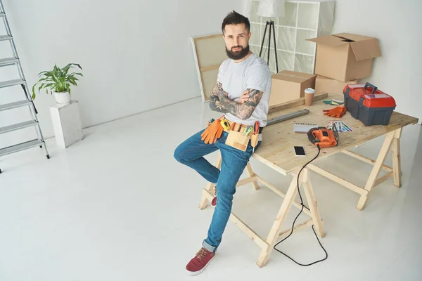 Vue grand angle du bel homme tatoué barbu avec ceinture à outils assis avec les bras croisés et regardant la caméra — Photo de stock