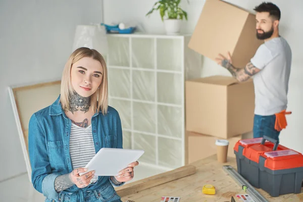 Young woman using digital tablet and looking at camera while boyfriend holding cardboard box behind — Stock Photo