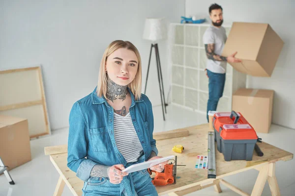 Chica sosteniendo la tableta digital y mirando a la cámara mientras el novio sostiene la caja de cartón detrás - foto de stock