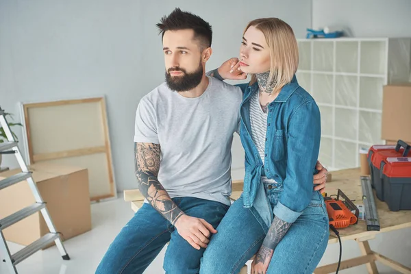 Young couple with tattoos sitting together and looking away in new home — Stock Photo