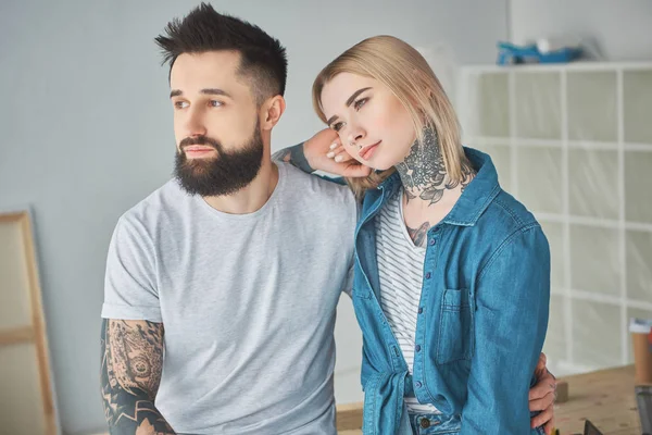 Belo jovem casal com tatuagens sentadas juntas e olhando para longe na nova casa — Fotografia de Stock