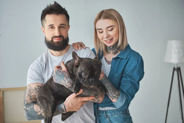 Feliz jovem casal com tatuagens segurando bonito bulldog francês em nova casa — Fotografia de Stock