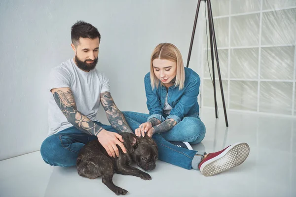 Feliz joven pareja sentado y jugando con bulldog francés en nueva casa - foto de stock