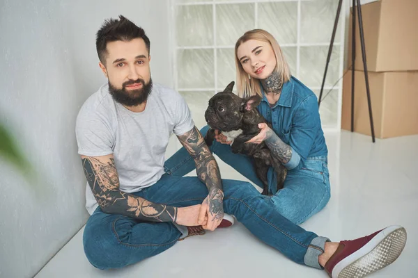 Happy young tattooed couple playing with french bulldog and smiling at camera in new home — Stock Photo