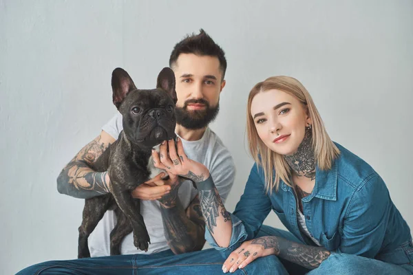 Jovem casal tatuado segurando bulldog francês e sorrindo para a câmera no cinza — Fotografia de Stock