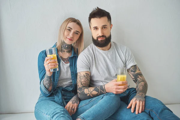 Joven pareja tatuada sosteniendo vasos de jugo y mirando a la cámara mientras está sentado en el suelo en casa nueva - foto de stock