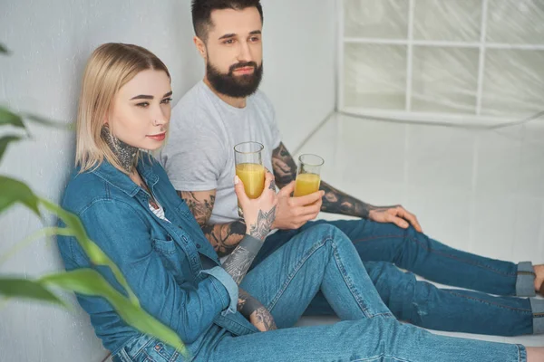 Young couple holding glasses of juice and looking away while sitting in floor in new house — Stock Photo