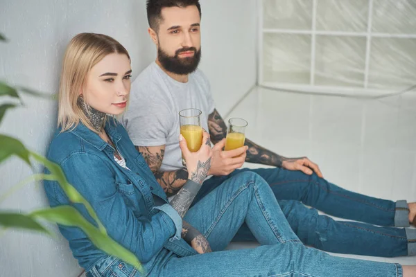 Pareja joven sosteniendo vasos de jugo y mirando hacia otro lado mientras se sienta en el piso en una casa nueva - foto de stock